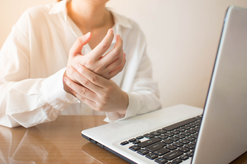 Photo of a person sitting in front of a laptop and holding on hand in the other, indicating soreness.