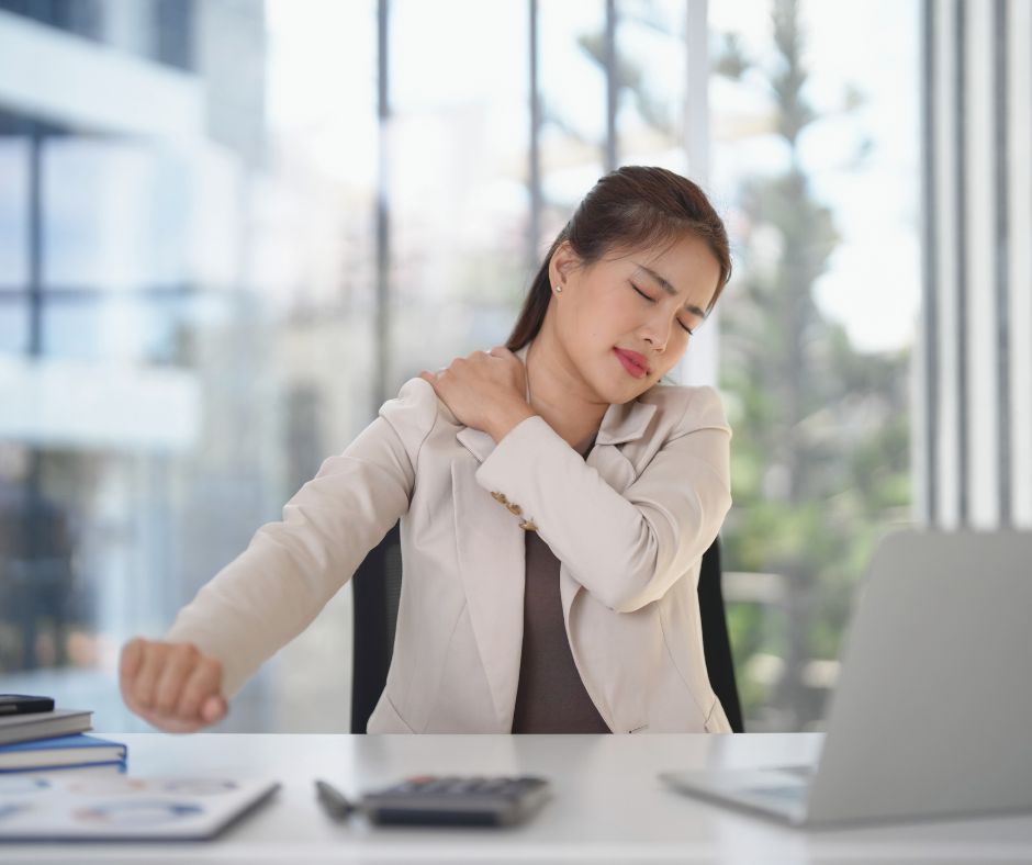 Photo of a woman sitting at a desk and holding her shoulder while grimacing in pain.
